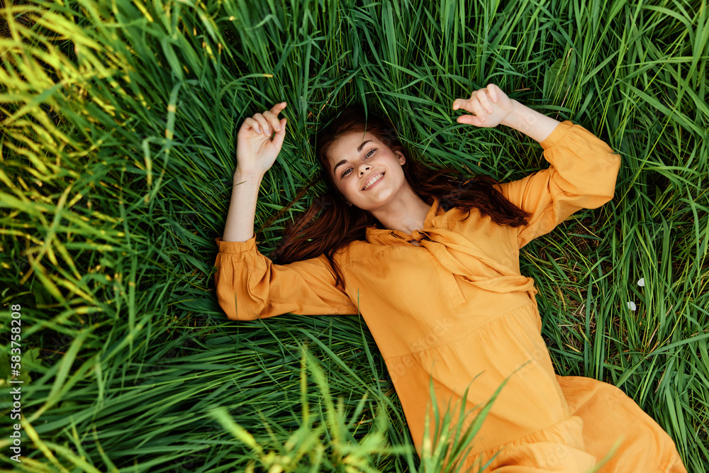 a joyful woman in a long orange dress lies in the tall green grass and looking at the camera smiles broadly