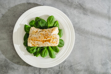 Wall Mural - Cod fillet baked with garlic butter sauce served with fresh spinach close-up on a white plate, flat lay