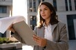 © opolja - Elegant young woman stands outdoors against the background of the business center, holding a folder with documents