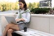 © opolja - Young pretty business woman working on laptop outside near modern building, sitting on remote work, drinking coffee