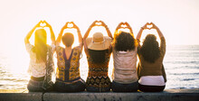 Group Of Women Outdoor Enjoying Leisure And Travel Summer Holiday Together In Friendship And Love Sitting Against A Sunset On The Ocean And Doing Heart Sig With Raised Up Arms And Hands. Beach Day