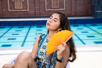 Exhausted Indian woman using a paper hand waver fan sitting outdoor in sunny day suffering from hot summer weather or high temperature, Beautiful asian Girl get uncomfortable due to heat wave.