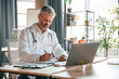 © standret - Sitting by table with laptop, writing data into document. Senior doctor in white coat is at work indoors