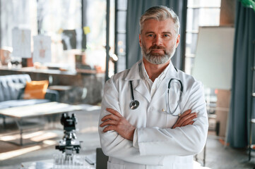 Wall Mural - Standing with arms crossed. Senior doctor in white coat is at work indoors