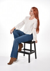 Wall Mural - full length portrait of beautiful woman model with long red hair, wearing casual outfit white blouse  top and denim jeans, isolated on white studio background. Relaxed seated pose, sitting on chair wi