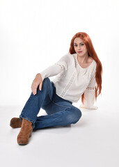 Canvas Print - full length portrait of beautiful woman model with long red hair, wearing casual outfit white blouse  top and denim jeans, isolated on white studio background. Relaxed seated pose, sitting on floor wi