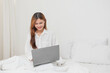 © PHAISITSAWAN - Woman in white nightgown waking up on weekend morning resting and relaxing playing with laptop mobile phone Eating bread and drinking tea in glass inside white bedroom.  Morning vacation concept.