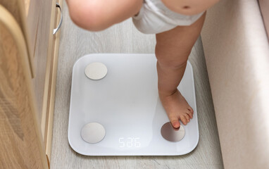 baby legs barefoot feet on body composition scale measuring weight home interior laminate wooden floor background bed and toddler crib.cute pretty fat infant legs on white scale digital numbers