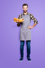 Canvas Print - Full size body portrait of positive attractive baker in jeans, shoes, shirt, apron with stubble having tray with bakery products, looking at camera, isolated on grey background