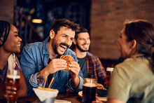 Happy Man Eats Burger While Talking To His Friends In Pub.