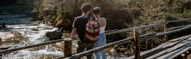 Wall Mural - Back view on couple of young hikers standing on wooden bridge over fast stream and hugging.