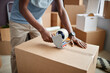 © Seventyfour - Close-up of African American man using adhesive tape to pack things into cardboard boxes for relocation