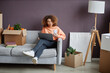 © Seventyfour - Young woman connecting her smartphone to laptop while sitting on the sofa among boxes after relocation