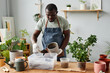 © Seventyfour - Waist up portrait of black man repotting plants indoors and scooping fresh soil mix, copy space