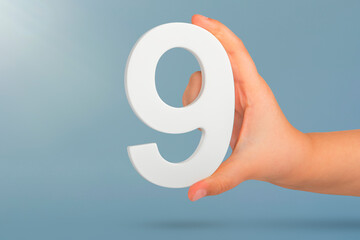 Fototapete - Number nine in hand. A hand holds a white number 9 on a blue background. Concept with number nine. Birthday 9 years, percentage, ninth grade or day.
