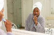 © Wavebreak Media - African american senior woman wearing bathrobe brushing teeth while looking in mirror in bathroom