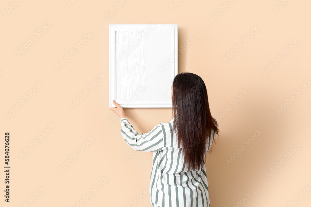 Young woman hanging blank picture on beige wall