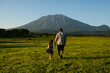 © Evgenii - Father and daughter are walking on the lawn, holding hands, rear view, against the backdrop of a beautiful mountain.