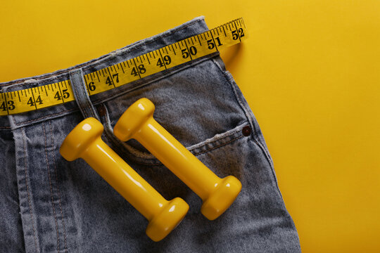 Wall Mural - Jeans with measuring tape and dumbbells on yellow background, top view. Weight loss concept