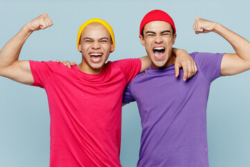 Young strong couple two friends men wear casual clothes looking camera together showing biceps muscles on hand demonstrating strength power isolated on pastel plain light blue cyan background studio.