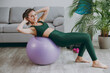 © ViDi Studio - Side view young strong sporty athletic fitness trainer instructor woman wear green tracksuit using lean back on fit ball training do abs exercises at home gym indoor. Workout sport motivation concept.