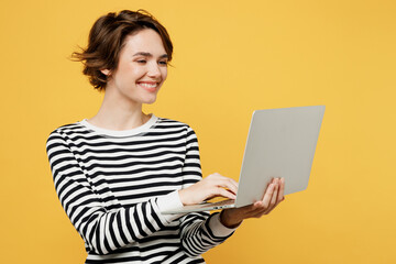 Wall Mural - Young smiling happy fun cheerful IT woman wears casual striped black and white shirt hold use work on laptop pc computer isolated on plain yellow color background studio portrait. Lifestyle concept.