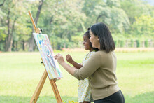 Asian Mother And Cute African Daughter Smiling Holding Same Paintbrush And Painting Coloring Watercolor On Canvas Together In Green Park On Summer. Mom Support Little Artist Girl In Outdoor Activity