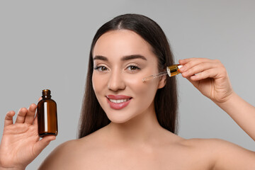 Wall Mural - Happy young woman with bottle applying essential oil onto face on light grey background