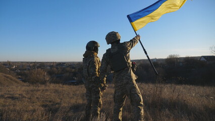 Dolly shot of young woman and man in military uniform waving flag of Ukraine against sunset. Female and male soldier of ukrainian army lifted blue-yellow banner holding hands of each other at field