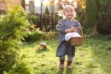 Happy little girl holding wicker basket with cute rabbit outdoors on sunny day