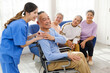 © artitwpd - The caregiver therapist stands with an Asian senior sitting in a wheelchair with a group of senior women sitting on a sofa in the background. The nursing home facilitates a support group.