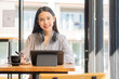 © ArLawKa - Asian woman relaxing hands using tablet surfing internet writing diary smiling cheerfully while sitting in cafe near large window