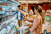 Little Girl Talks To Her Mother While Shopping In Supermarket And Pointing At Product She Wants To Buy.