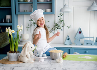 little funny cute smiling girl wearing a white chef's hat and uniform sitting near bowl of dough for easter bread in the blue and white kitchen and playing with toy rabbit