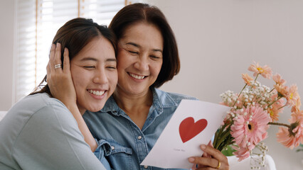 Wall Mural - May Mother's day young adult grown up child cuddle hug give flower gift box red heart card to mature middle aged mum. Love kiss care mom asia people sitting at home sofa happy smile enjoy family time.