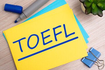 Poster - toefl. text on yellow paper on light wooden background with stationery