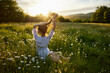 © VICHIZH - a red-haired woman in a light dress sits with her back to the camera in a field of daisies straightening her hair with her hands against the backdrop of the setting sun