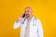 © Designerant - Telemedicine, close up portrait of middle aged bald doctor making telemedicine. Physician using smartphone. Isolated yellow background, copy space. Wearing white hospital coat, stethoscope.