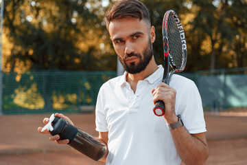 Holding bottle with water and racket. Young man is on the tennis court at sunny daytime