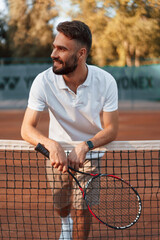 Wall Mural - Standing by the net with racket. Young man is on the tennis court at sunny daytime