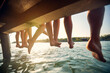 © luckybusiness - Rear view of legs of group of friends by lake. Four young friends sitting on jetty with their legs hanging down to the water on a summer day. Holiday, togetherness, lifestyle concept.