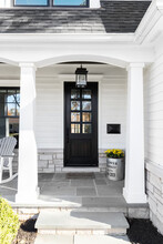 A New, White Modern Farmhouse With A Dark Wood Door With Windows, White Pillars, A Stone Floor, And Patio Furniture.