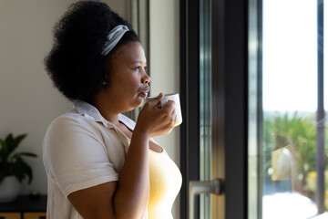 Thoughtful plus size african american woman looking through window and drinking coffee