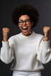 © Alexandre - Mulher afro-americana muito feliz cerra os punhos com triunfo, ri de felicidade, regozija-se com o sucesso, usa óculos, suéter branco, torce pelo time favorito, isolado em fundo cinza