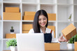 © wichayada - A portrait of a young Asian woman, e-commerce employee sitting in the office full of packages in the background write note of orders and a calculator, for SME business ecommerce and delivery business