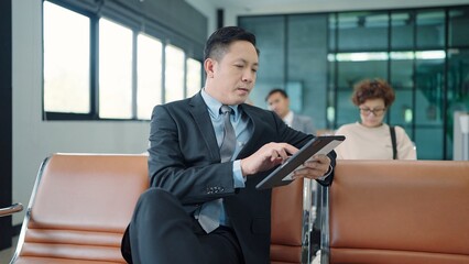 Asian businessman wearing suit sitting and working on tablet and waiting for flight at airport terminal