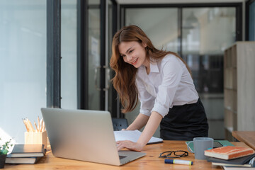 Attractive young Asian businesswoman standing leaning over her desk, concentrating on her project on laptop.