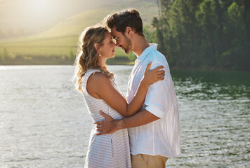 Couple, head touch and hug by lake for outdoor date, romantic adventure and summer love in nature. Man, woman and romance with embrace, care and bond by water with sunshine, freedom and happiness