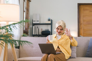 Wall Mural - Attractive and happy Muslim millennial businesswoman wearing a hijab sitting on a sofa in the living room, using a laptop computer for a video chat and waving.