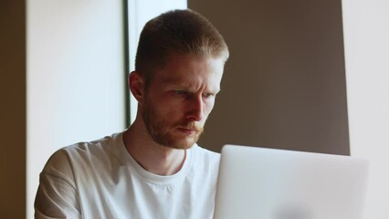 A concentrated man in a white T-shirt looks at the screen of his laptop. Tense man working on a laptop trying to finish a work project by a deadline.